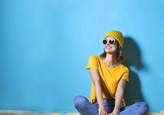 Fashionable young woman sitting indoors wearing sunglasses and a yellow top on a bright blue background.