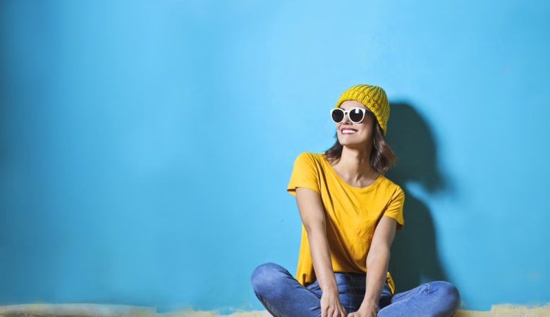 Fashionable young woman sitting indoors wearing sunglasses and a yellow top on a bright blue background.